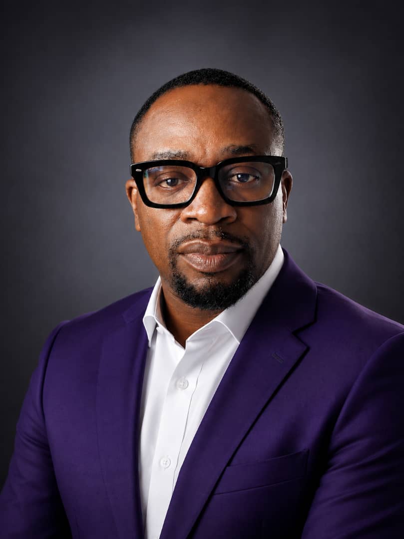 Mr. Jyno, a formal Black Nigerian teacher in a neutral classroom setting